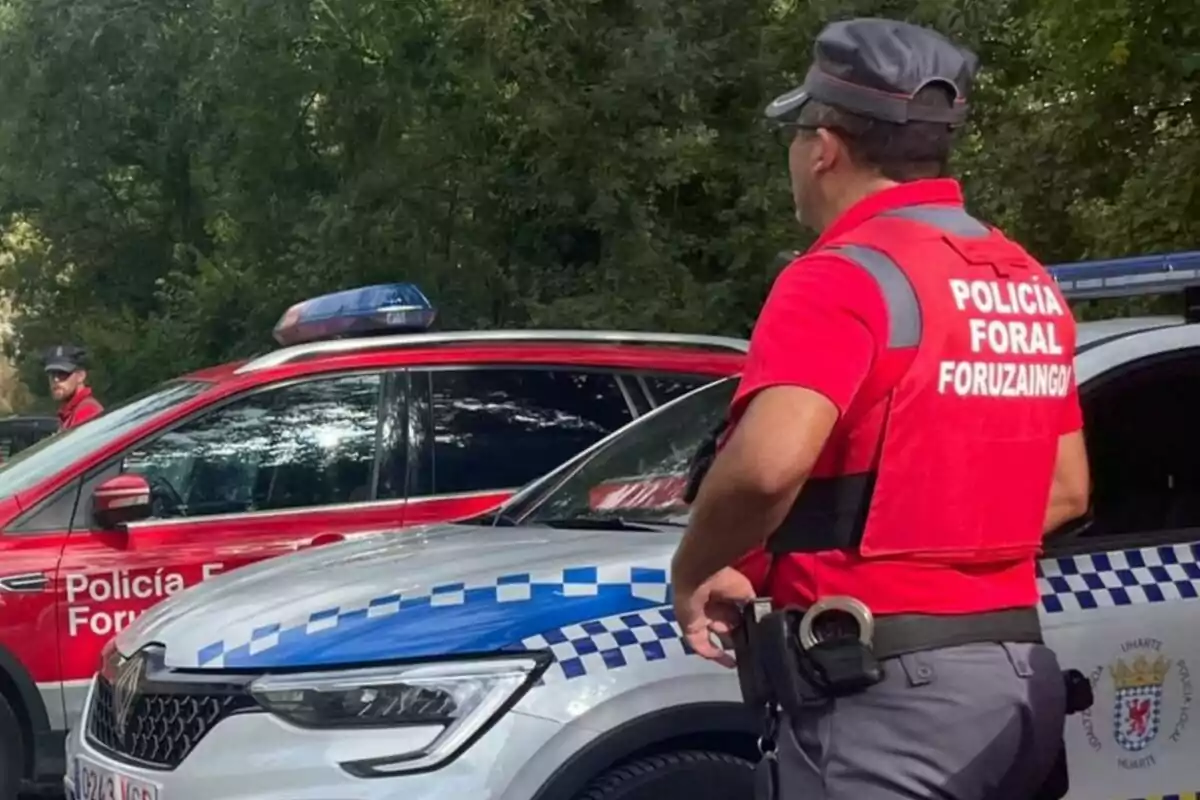 Agente de la Policía Foral de Navarra de espaldas junto a vehículos patrulla en una zona arbolada Agente de la Policía Foral de Navarra de espaldas junto a vehículos patrulla en una zona arbolada