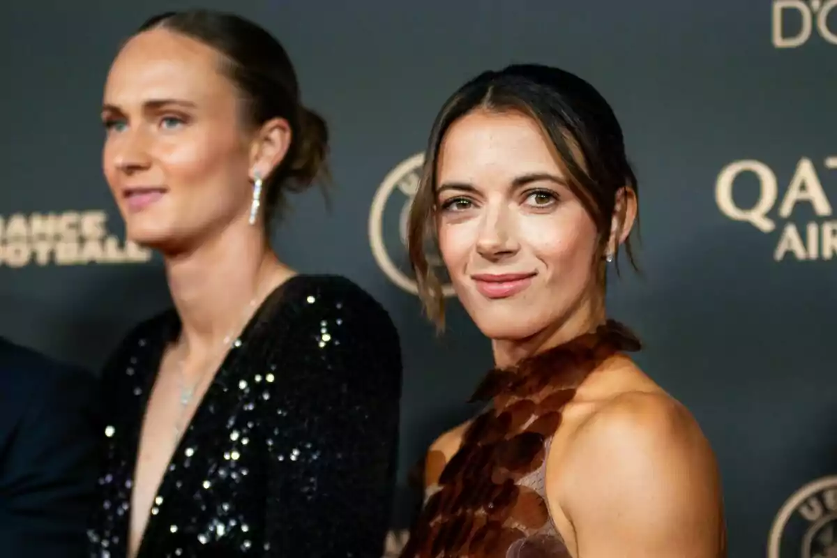 Dues dones elegantment vestides posen en una catifa vermella davant d’un fons amb logotips daurats.