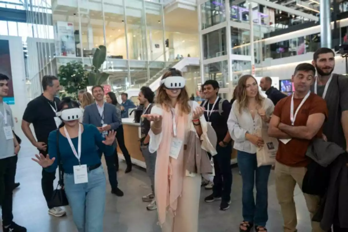 Personas reunidas en un espacio moderno mientras dos mujeres en el centro usan visores de realidad virtual y el resto observa con interés. Personas reunidas en un espacio moderno mientras dos mujeres en el centro usan visores de realidad virtual y el resto observa con interés.
