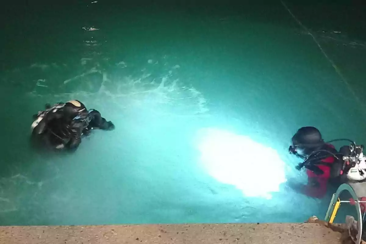 Dos buzos equipados con trajes de buceo y tanques de oxígeno se encuentran en el agua iluminada, uno de ellos sostiene una luz brillante bajo la superficie.