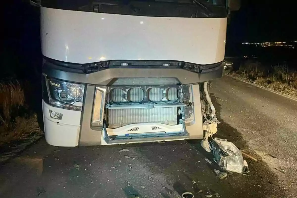 Camió blanc amb la part davantera danyada i peces despreses sobre una carretera de nit