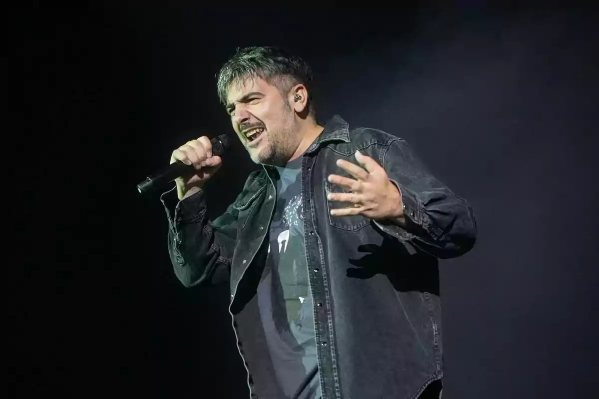 Home cantant apassionadament a l’escenari amb micròfon a la mà i jaqueta texana fosca Home cantant apassionadament a l’escenari amb micròfon a la mà i jaqueta texana fosca