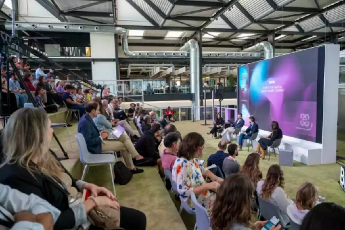 Personas sentadas en gradas y sillas asisten a una conferencia en un espacio moderno con un panel de expertos frente a una pantalla grande. Personas sentadas en gradas y sillas asisten a una conferencia en un espacio moderno con un panel de expertos frente a una pantalla grande.