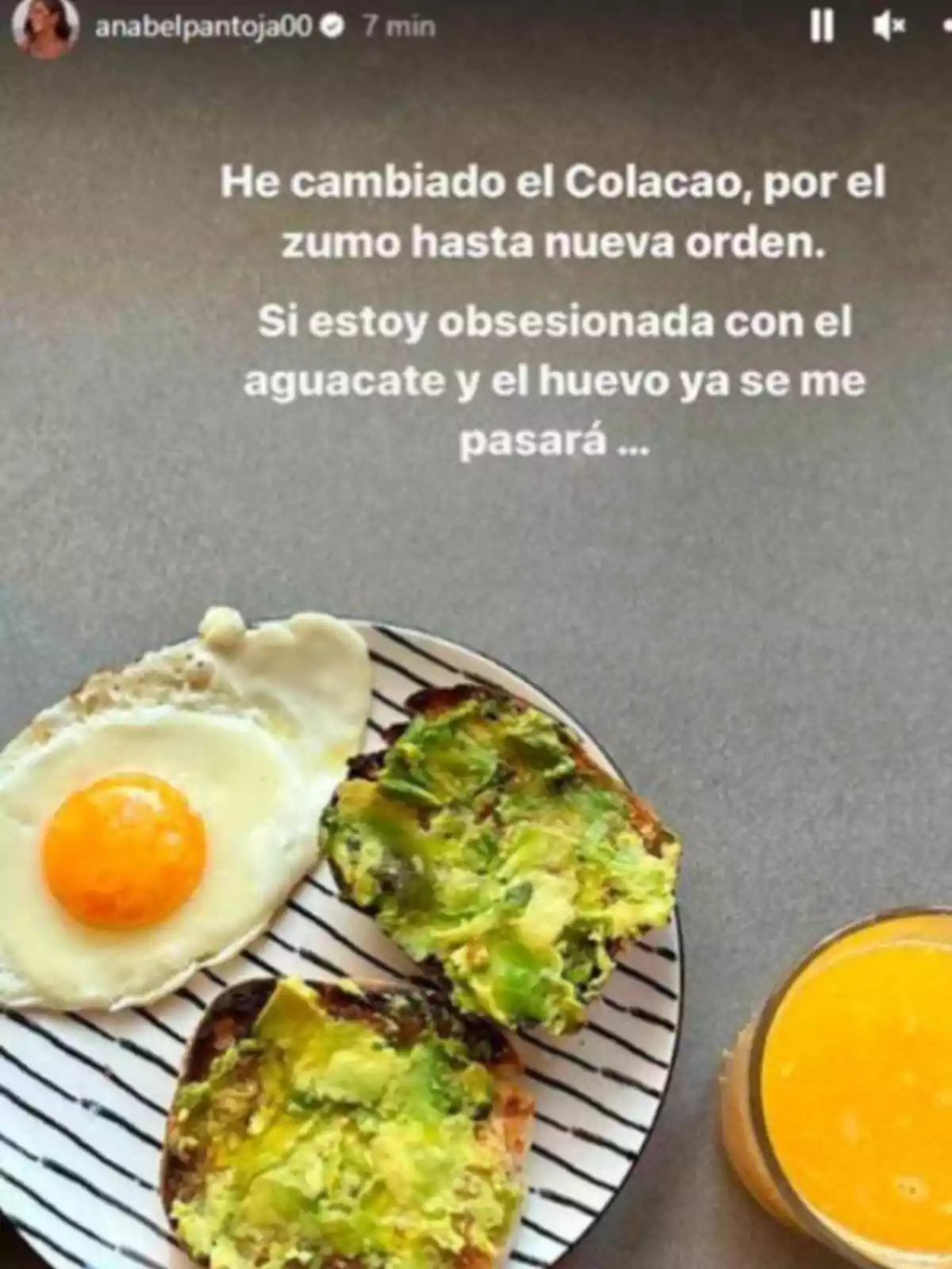 Desayuno con huevo frito, pan con aguacate y un vaso de zumo de naranja sobre una mesa gris