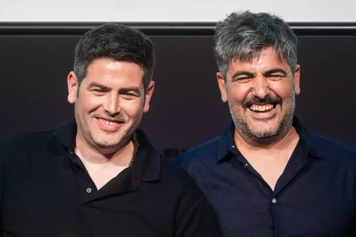 Dos homes somriuen i posen junts davant de la càmera, tots dos porten camises fosques i es veuen feliços. Dos homes somriuen i posen junts davant de la càmera, tots dos porten camises fosques i es veuen feliços.