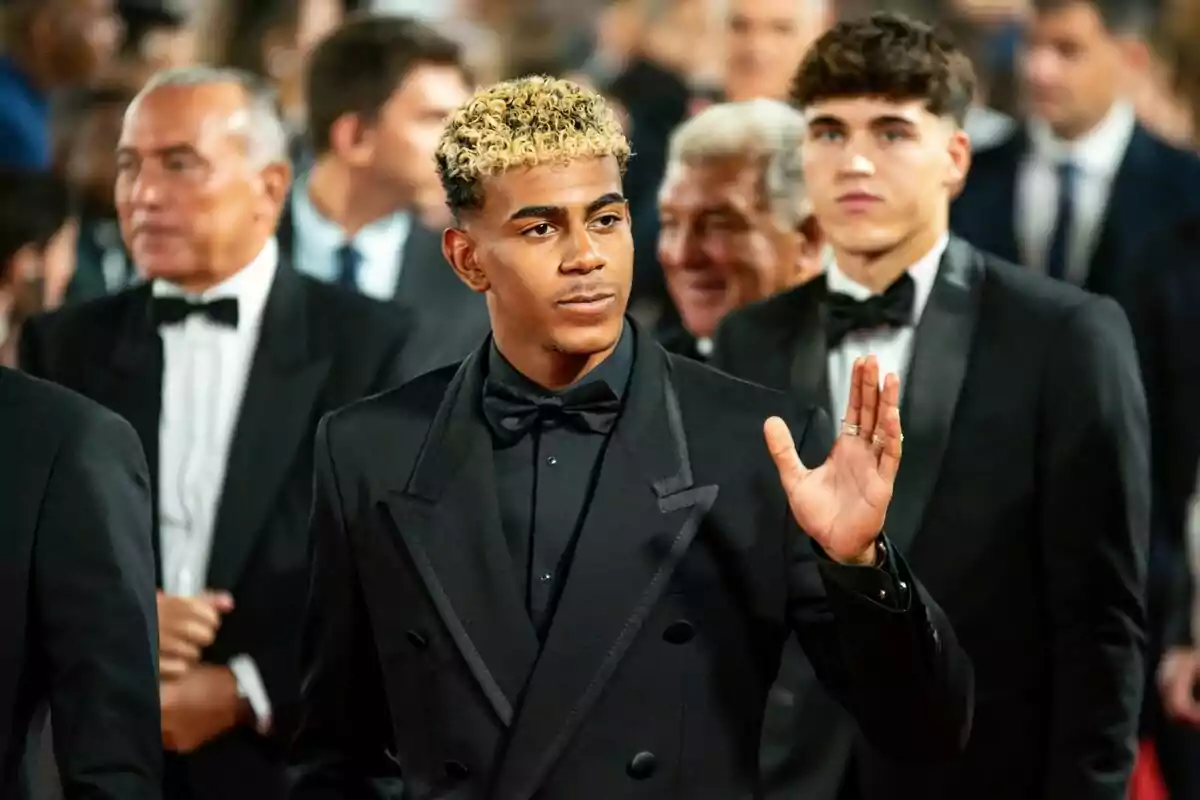 Jove amb cabells arrissats rossos i vestit negre saluda mentre camina entre una multitud de persones vestides de gala. Jove amb cabells arrissats rossos i vestit negre saluda mentre camina entre una multitud de persones vestides de gala.