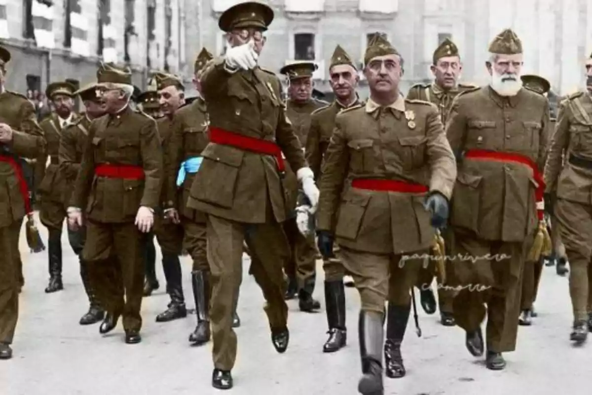 Un grup de militars amb uniformes marrons i cinturons vermells marxa per un carrer urbà mentre un d’ells assenyala cap endavant.