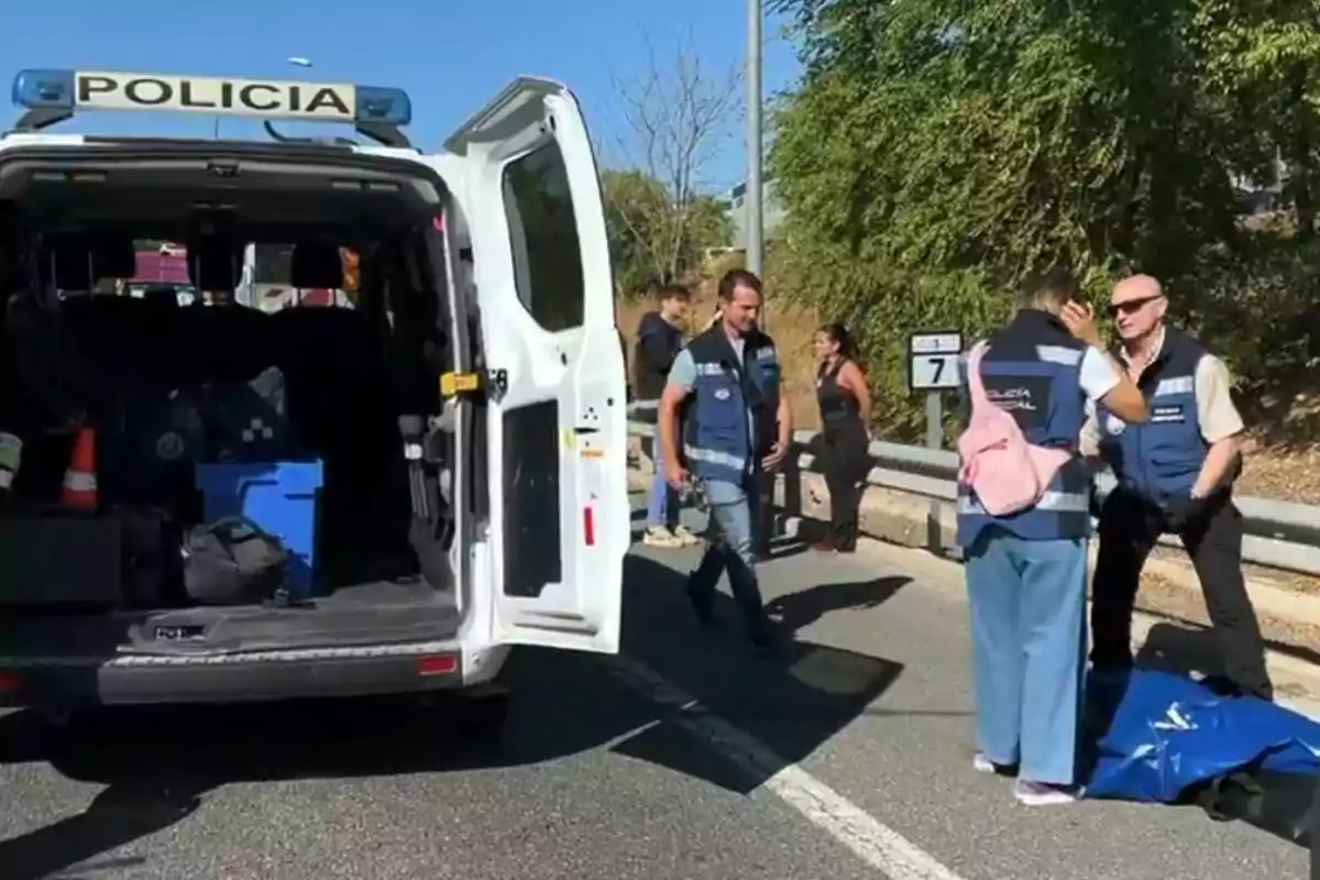 Diversos agents de policia investiguen una escena en una carretera al costat d’una furgoneta policial amb la porta del darrere oberta i un cos cobert a terra