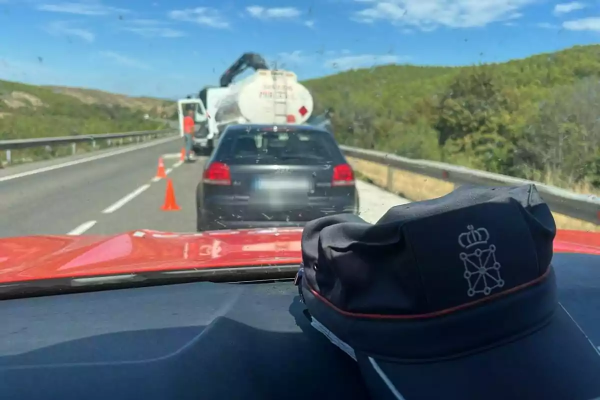 Un coche detenido en una carretera detrás de un camión cisterna con conos naranjas y un gorro de policía foral sobre el tablero del vehículo Un coche detenido en una carretera detrás de un camión cisterna con conos naranjas y un gorro de policía foral sobre el tablero del vehículo