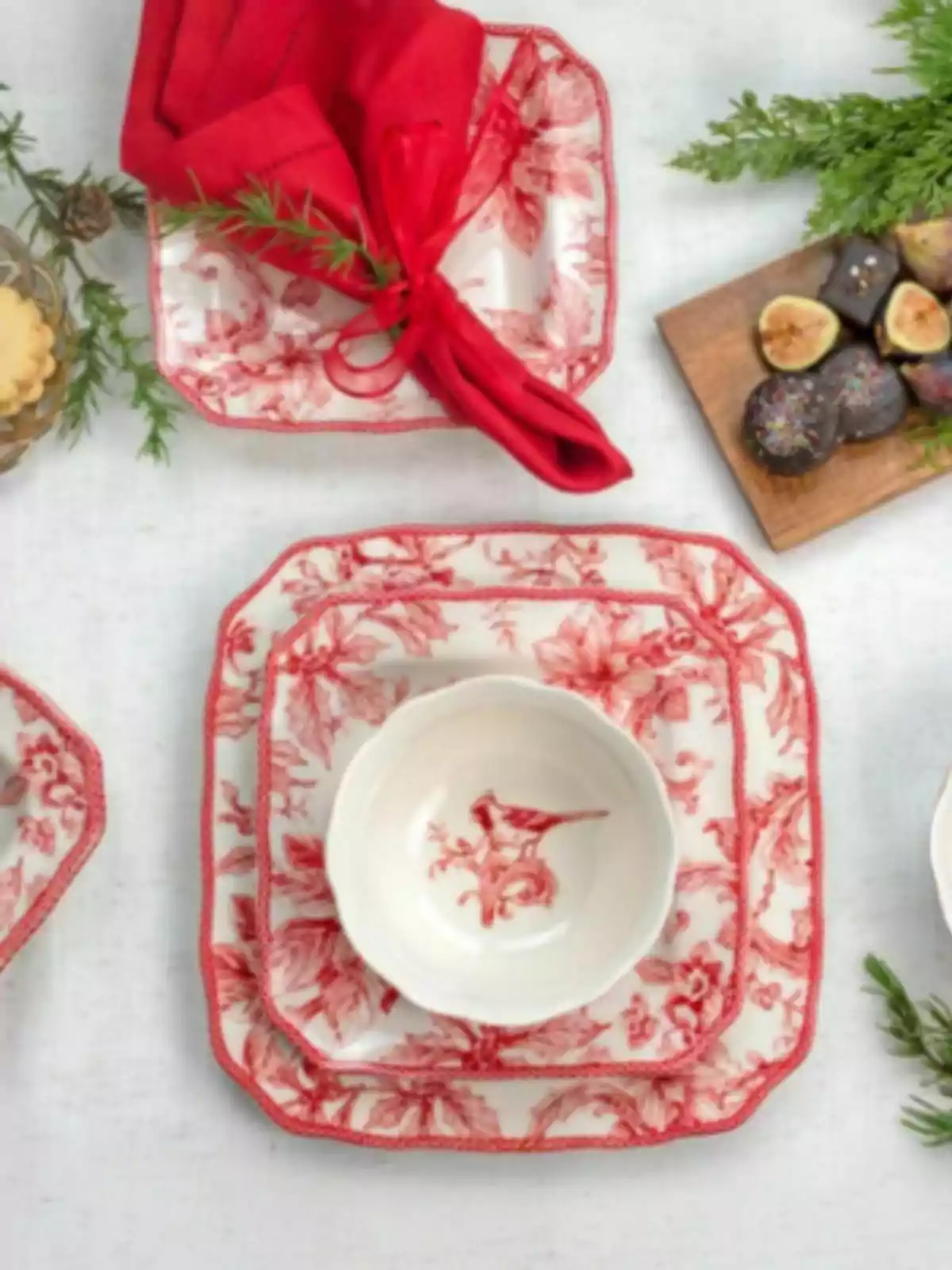 Vajilla blanca con detalles rojos de estilo navideño, servilleta roja atada con lazo y ramita de romero, acompañada de postres y decoraciones naturales. Vajilla blanca con detalles rojos de estilo navideño, servilleta roja atada con lazo y ramita de romero, acompañada de postres y decoraciones naturales.