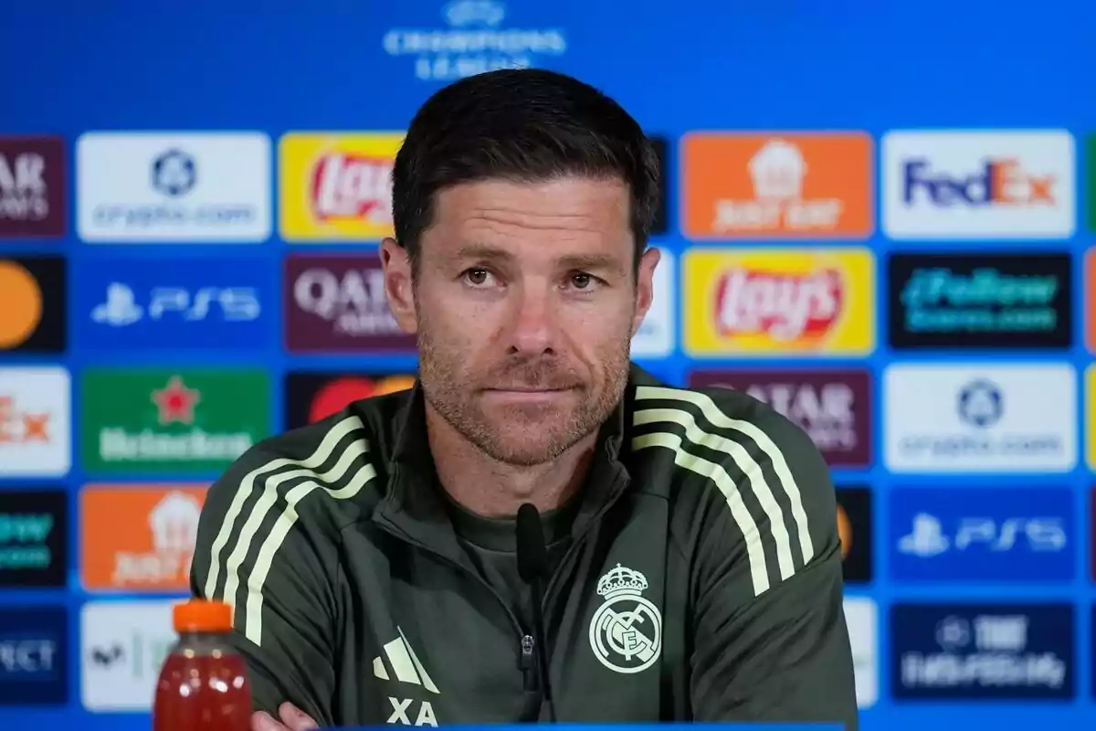 Home amb jaqueta esportiva del Reial Madrid assegut davant d’un micròfon en una roda de premsa amb fons de patrocinadors de la Lliga de Campions Home amb jaqueta esportiva del Reial Madrid assegut davant d’un micròfon en una roda de premsa amb fons de patrocinadors de la Lliga de Campions