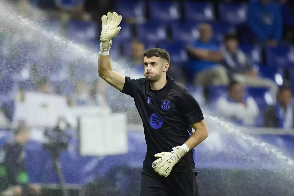 Portero con uniforme negro y guantes levantando la mano en un estadio mientras el sistema de riego está encendido Portero con uniforme negro y guantes levantando la mano en un estadio mientras el sistema de riego está encendido