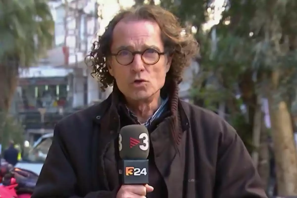 Un reporter amb ulleres i cabells arrissats, que és Joan Roura, sosté un micròfon de TV3 mentre informa des d’un carrer a l’aire lliure. Un reporter amb ulleres i cabells arrissats, que és Joan Roura, sosté un micròfon de TV3 mentre informa des d’un carrer a l’aire lliure.