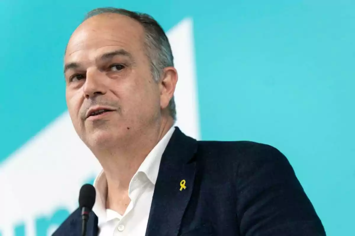 Home de mitjana edat amb jaqueta fosca i camisa blanca parlant davant d’un micròfon sobre un fons blau turquesa Home de mitjana edat amb jaqueta fosca i camisa blanca parlant davant d’un micròfon sobre un fons blau turquesa