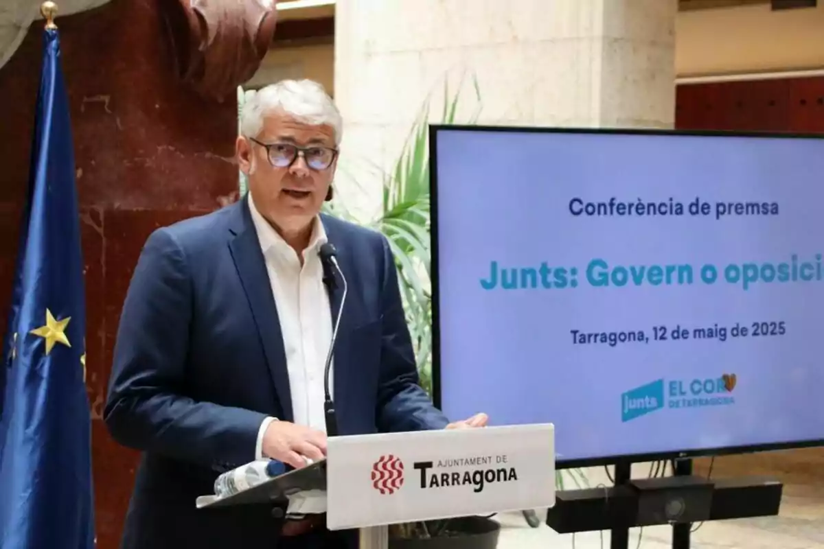 Un hombre de cabello canoso y gafas habla en una rueda de prensa frente a un atril del Ayuntamiento de Tarragona, con una pantalla a su lado que muestra información sobre una conferencia titulada Junts Govern o oposició en Tarragona el 12 de mayo de 2025.