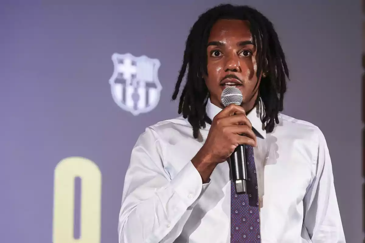 Joules Koundé amb camisa blanca i corbata parlant per micròfon davant d’un fons amb l’escut del FC Barcelona.