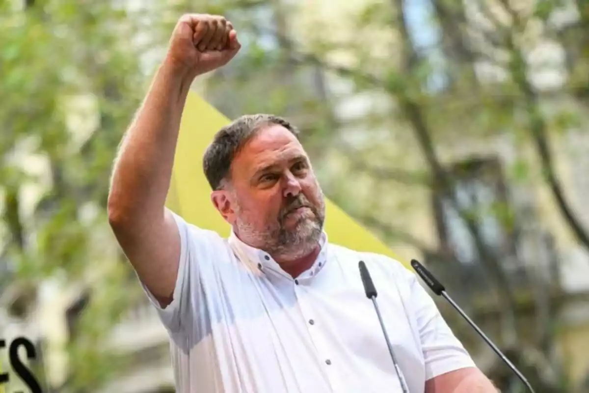 Hombre de barba y camisa blanca levantando el puño derecho mientras habla frente a dos micrófonos en un entorno al aire libre con árboles y edificios de fondo