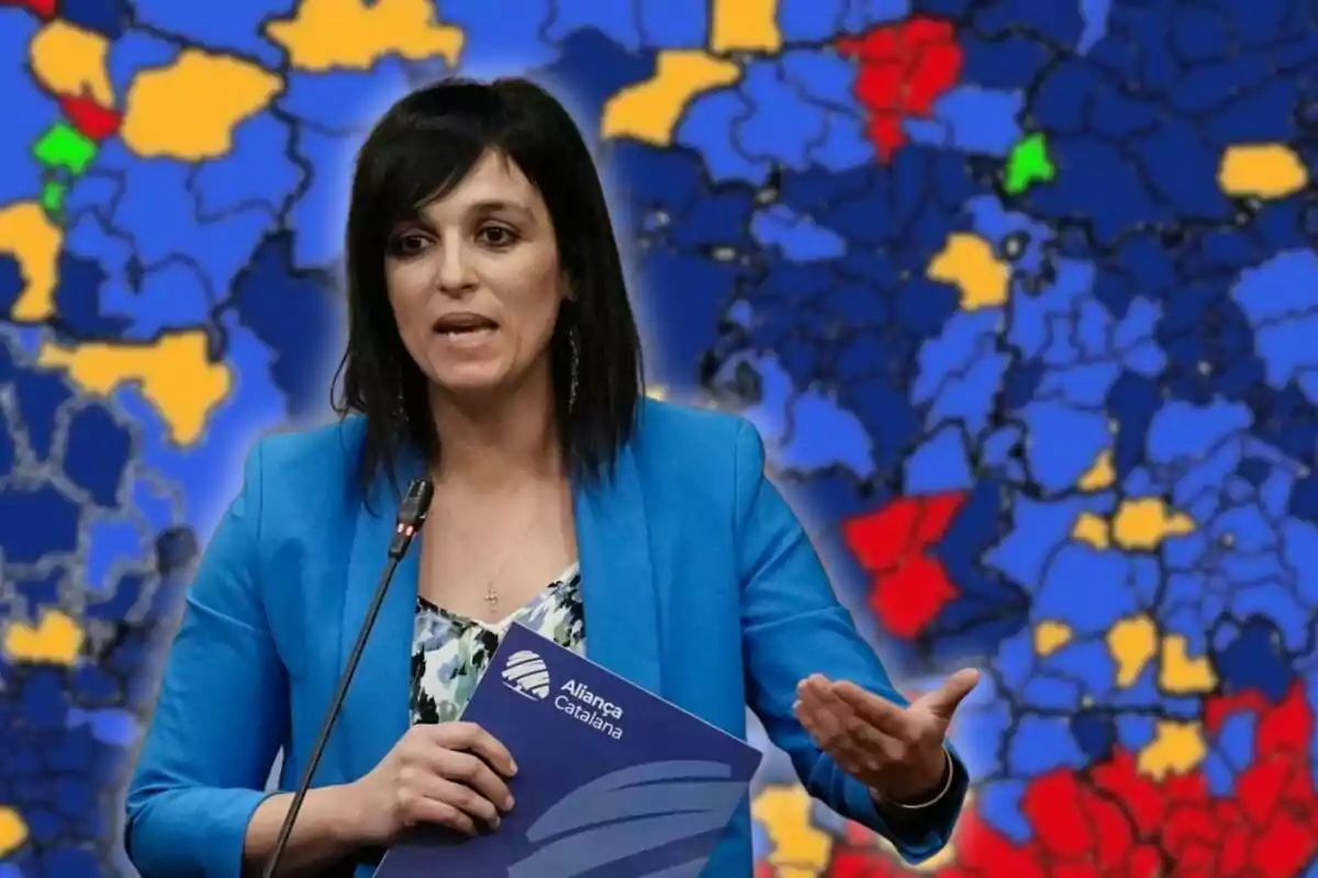 Dona de cabell fosc amb jaqueta blava parlant davant d’un micròfon i sostenint una carpeta d’Aliança Catalana amb un mapa polític de fons en colors blau, groc, vermell i verd