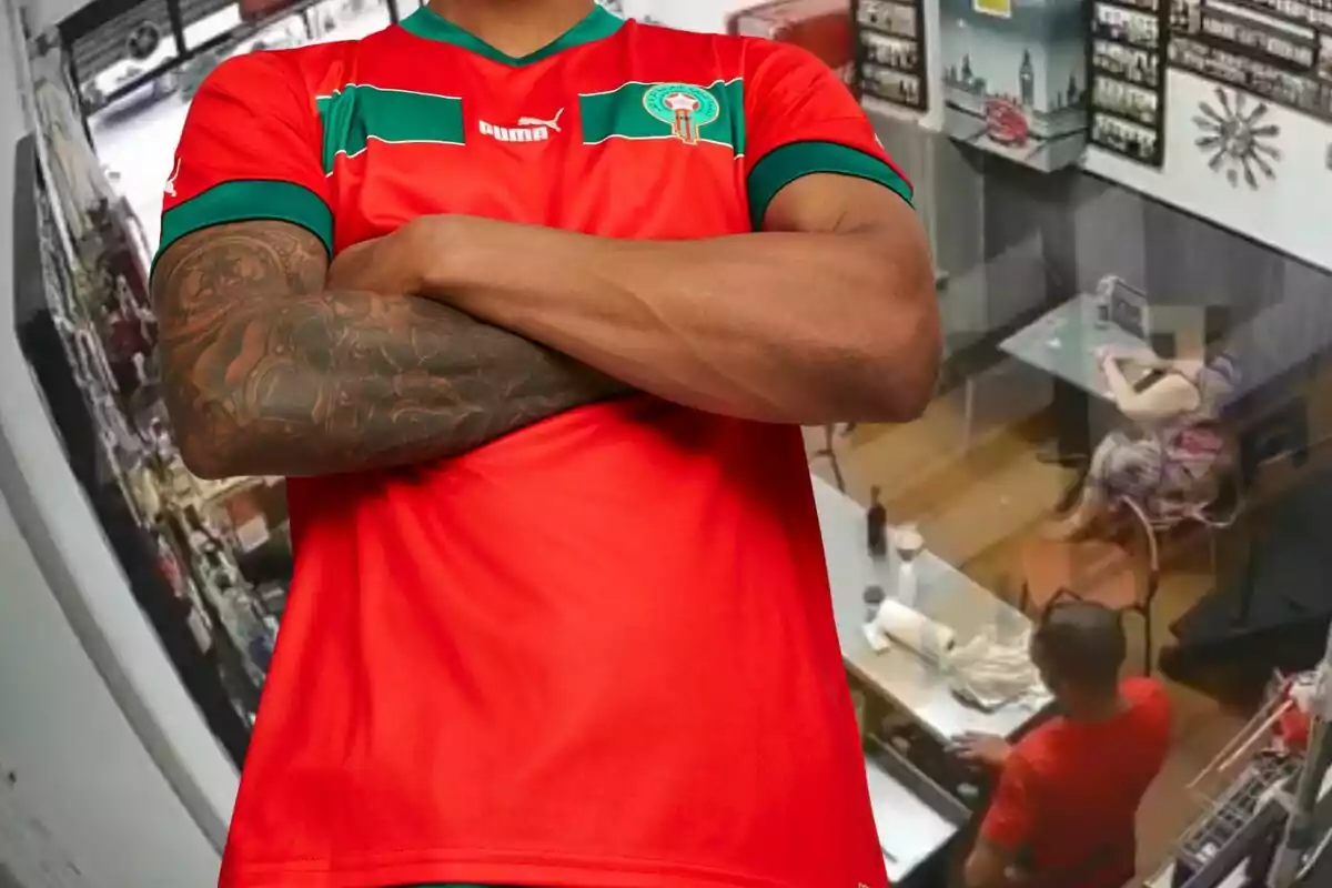 Persona amb samarreta vermella i verda de futbol amb els braços creuats en primer pla i al fons una escena de restaurant amb una dona asseguda i un home dret al costat d’una taula.