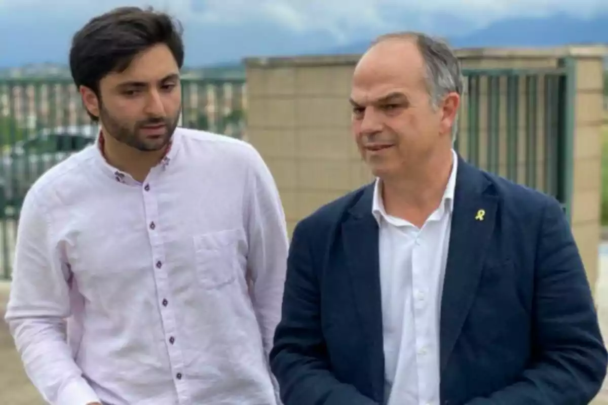 Dos hombres conversan al aire libre, uno lleva camisa clara y el otro viste saco azul marino con una camisa blanca