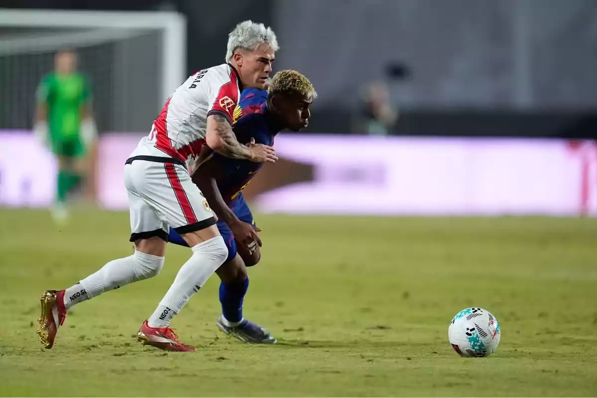 Dos futbolistas disputan el balón durante un partido en un estadio, uno con uniforme blanco y rojo y el otro con uniforme azul oscuro Dos futbolistas disputan el balón durante un partido en un estadio, uno con uniforme blanco y rojo y el otro con uniforme azul oscuro