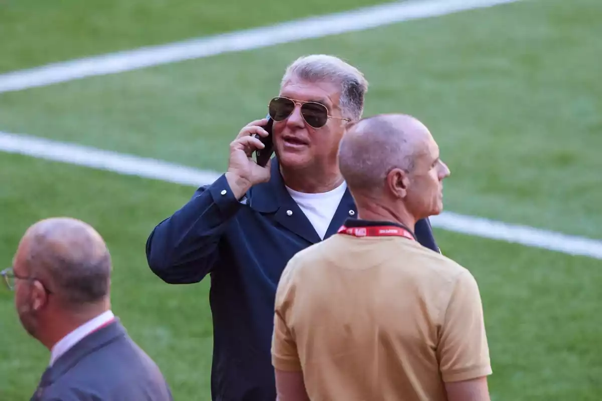 Hombre con gafas de sol hablando por teléfono en un campo de fútbol acompañado de otras dos personas Hombre con gafas de sol hablando por teléfono en un campo de fútbol acompañado de otras dos personas