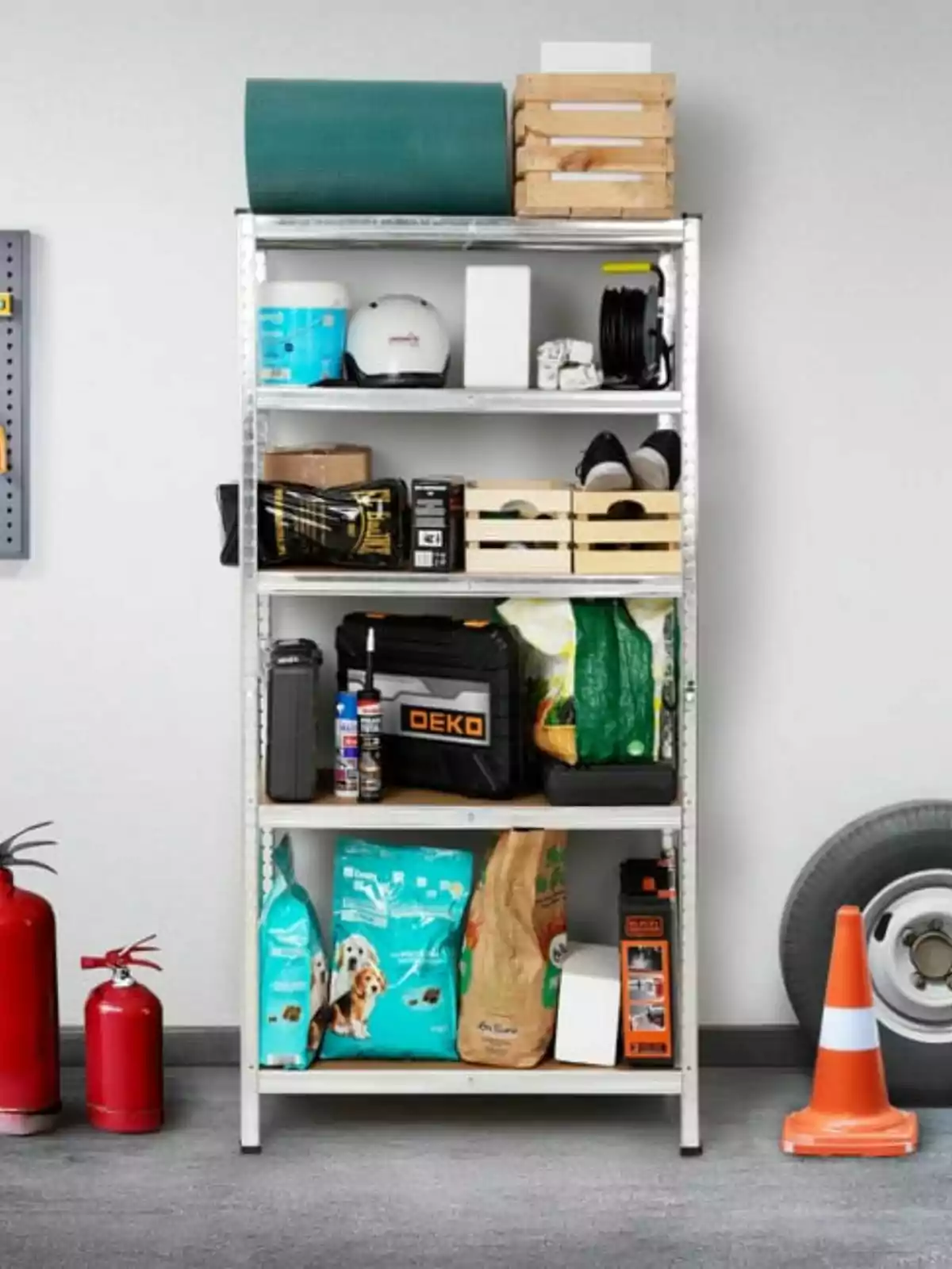 Estante metálico con varios objetos organizados como cajas, herramientas, bolsas de alimento para mascotas y equipo de seguridad, junto a extintores, una llanta y un cono de tráfico en un garaje.