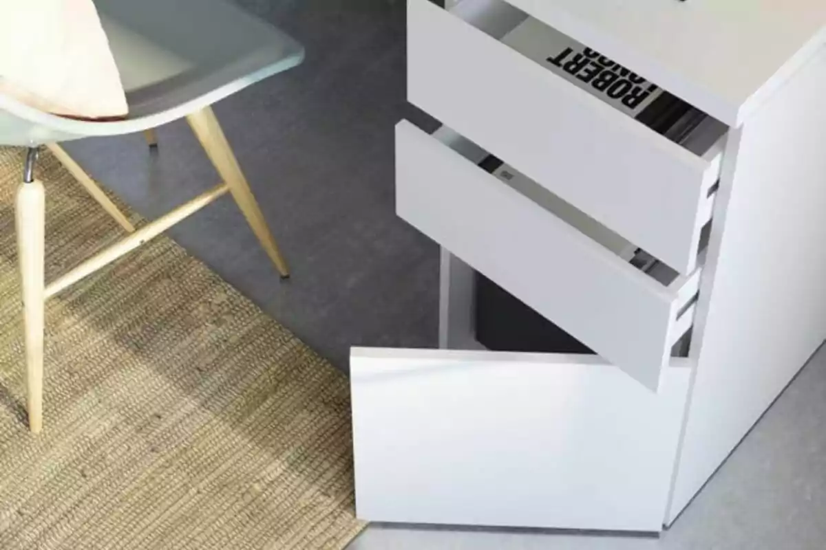 Cajonera blanca con tres cajones abiertos junto a una silla gris y una alfombra tejida sobre piso gris