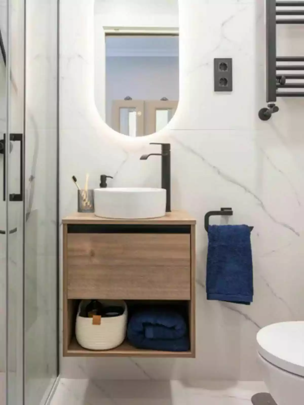 Baño moderno con lavabo sobre mueble de madera, espejo ovalado iluminado, toalla azul colgada y detalles en negro Baño moderno con lavabo sobre mueble de madera, espejo ovalado iluminado, toalla azul colgada y detalles en negro