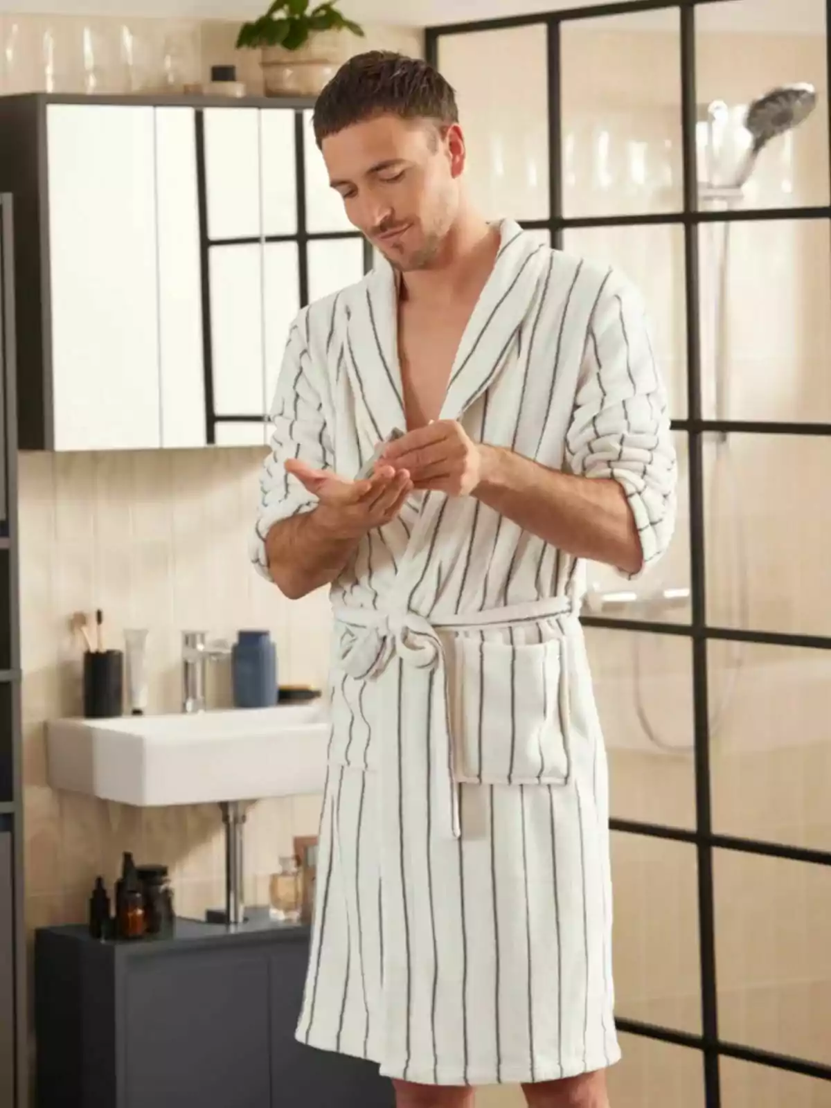 Home amb barnús blanc amb ratlles aplicant crema a les mans en un bany modern