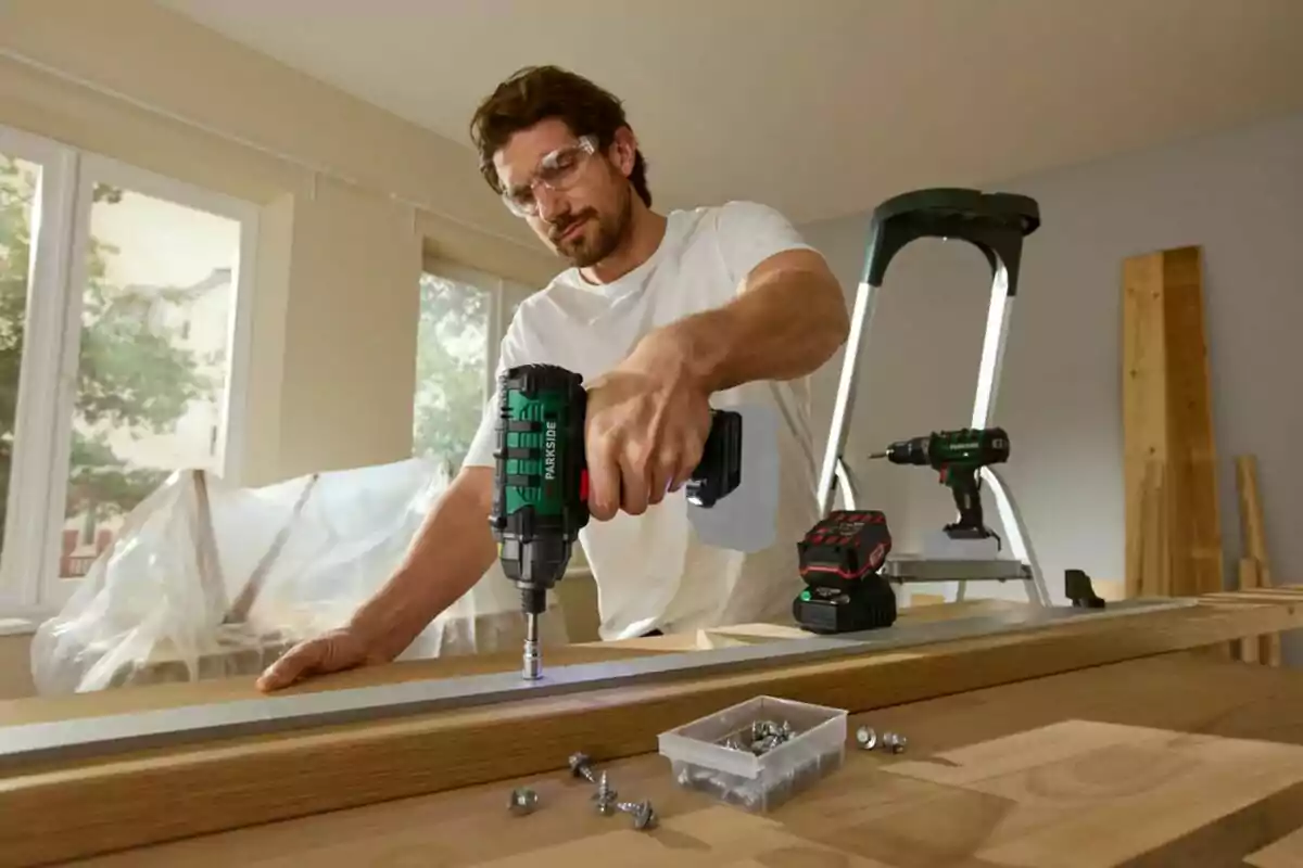 Man using an electric drill to screw a metal piece onto a wooden table in an indoor setting with tools and screws around.