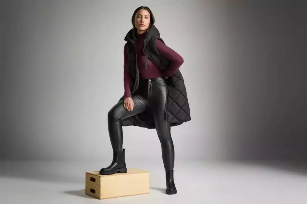 Mujer posando con chaleco acolchado negro, suéter burdeos, pantalones de cuero negro y botas negras sobre una caja de madera en un estudio con fondo gris Mujer posando con chaleco acolchado negro, suéter burdeos, pantalones de cuero negro y botas negras sobre una caja de madera en un estudio con fondo gris