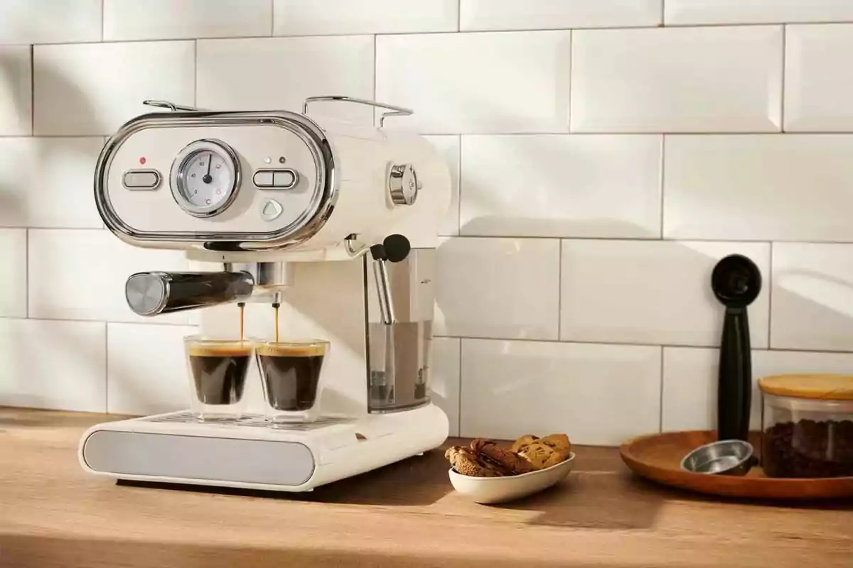 Cafetera blanca preparando dos espressos en una cocina con galletas y café en grano sobre la mesa Cafetera blanca preparando dos espressos en una cocina con galletas y café en grano sobre la mesa
