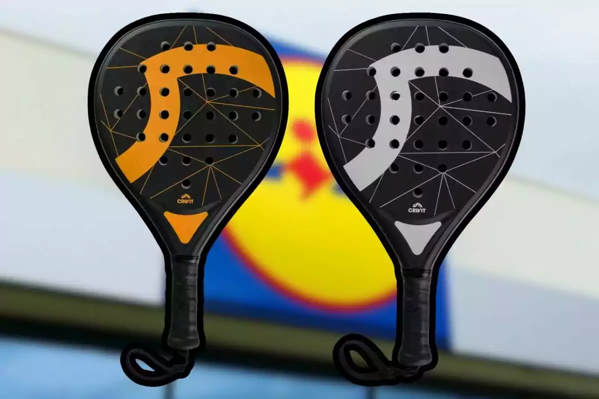 Dos palas de pádel negras con detalles en naranja y gris sobre un fondo desenfocado de un logotipo amarillo y azul