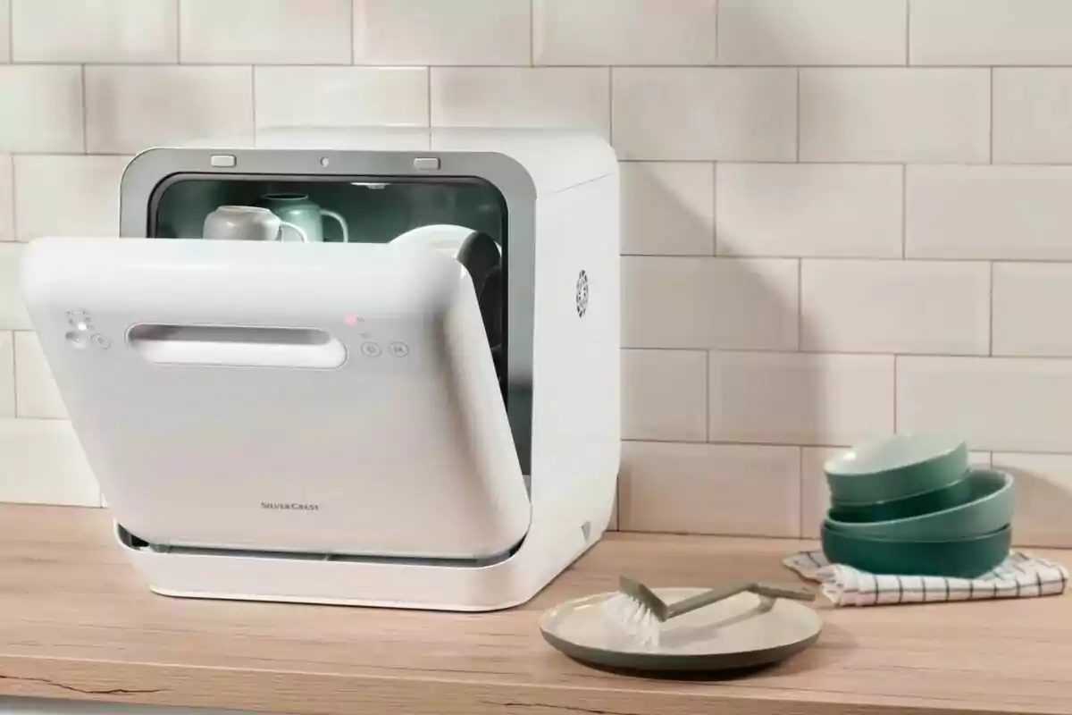 Rentavaixelles compacte de color blanc sobre una tauleta de fusta amb la porta oberta i alguns plats i tasses a l’interior, al costat d’un plat, un raspall i diversos bols apilats a un costat.