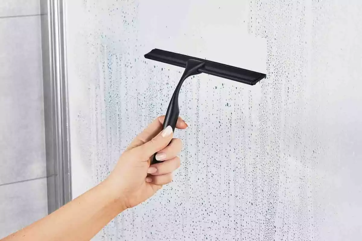 Mano usando un limpiavidrios negro para quitar el agua de una mampara de baño Mano usando un limpiavidrios negro para quitar el agua de una mampara de baño