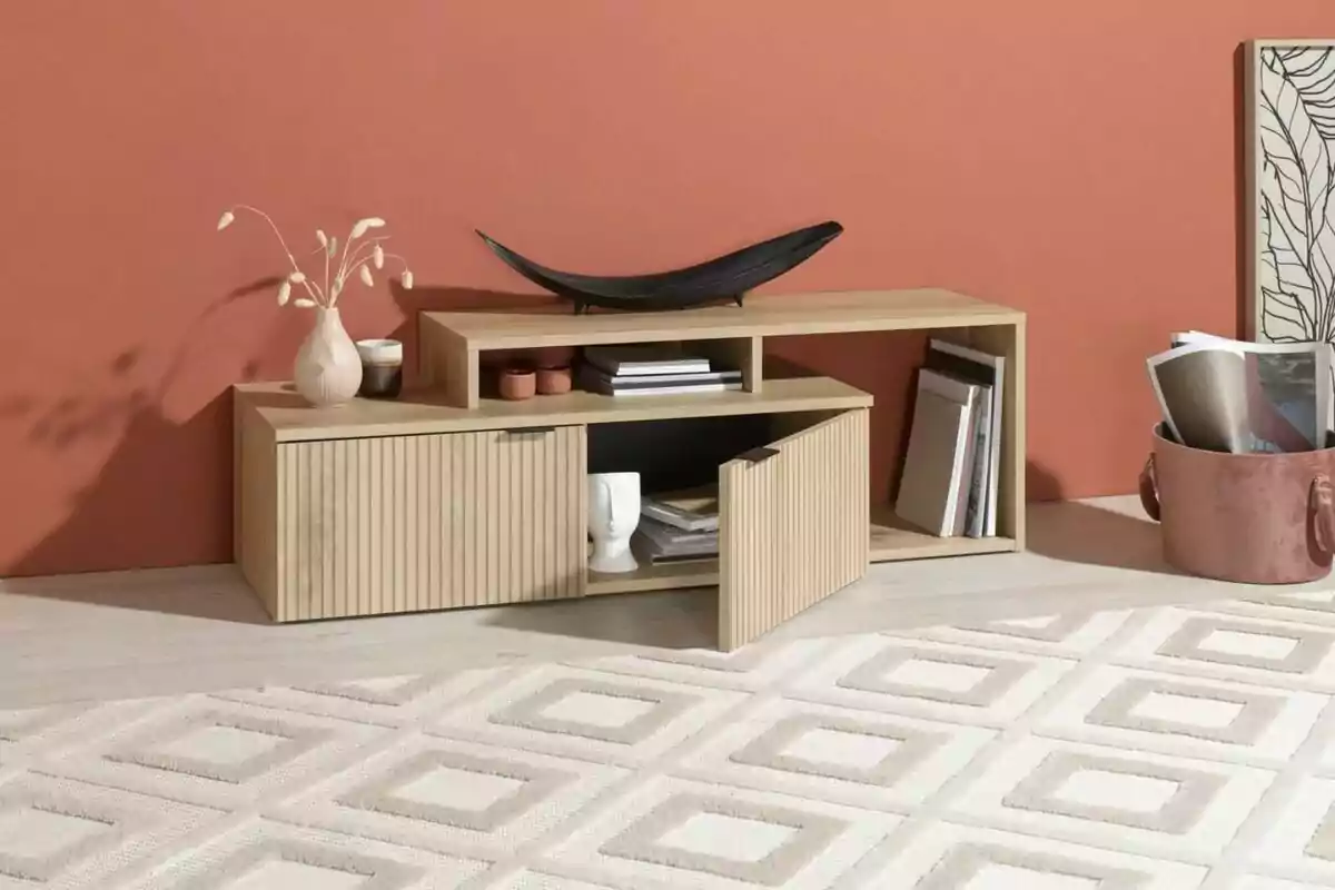 Mueble bajo de madera clara con puertas abiertas y estantes, decorado con jarrón, libros y objetos, sobre una alfombra clara con patrón geométrico y una pared color terracota de fondo