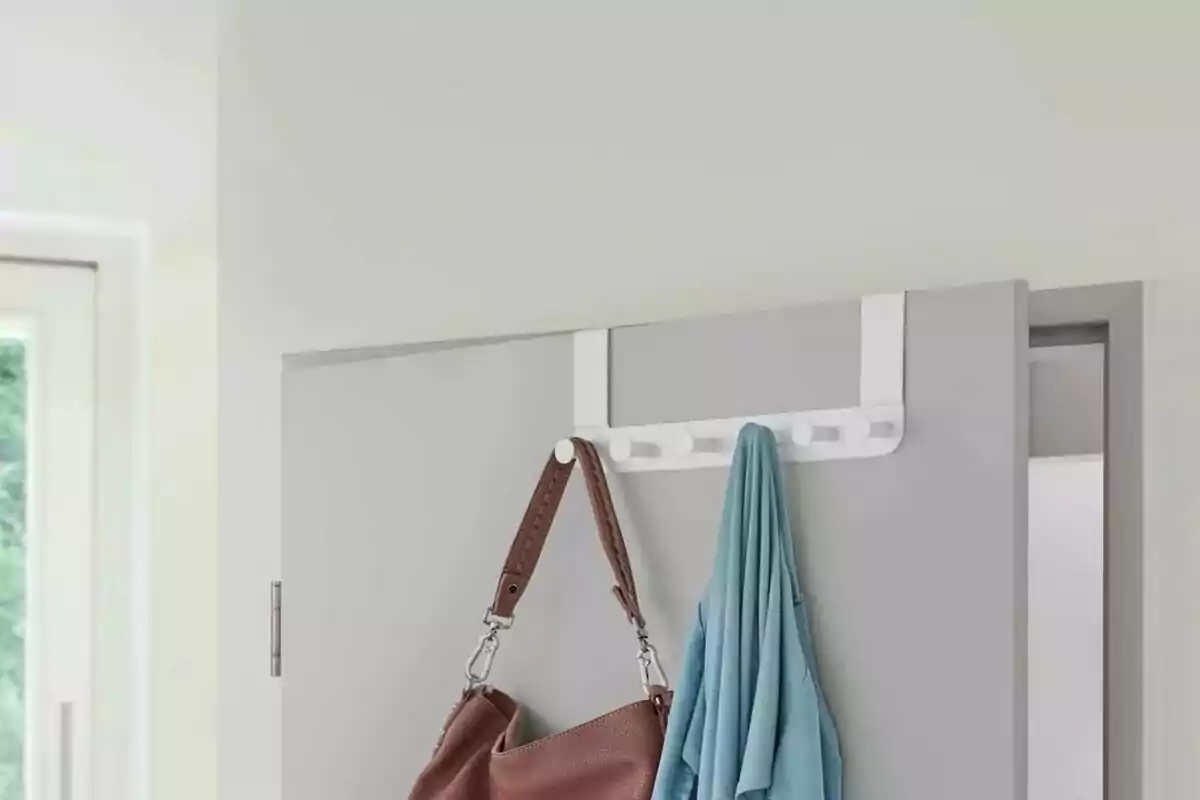 Perchero blanco colgado en la parte superior de una puerta gris con un bolso marrón y una prenda azul colgados Perchero blanco colgado en la parte superior de una puerta gris con un bolso marrón y una prenda azul colgados