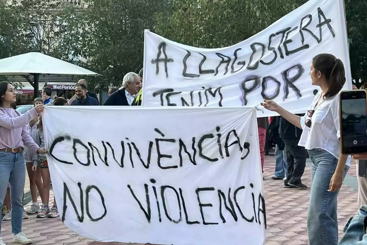 Persones sostenen pancartes en una manifestació, una d’elles diu convivència no violència i l’altra a Llagostera tenim por, en una plaça a l’aire lliure. Persones sostenen pancartes en una manifestació, una d’elles diu convivència no violència i l’altra a Llagostera tenim por, en una plaça a l’aire lliure.