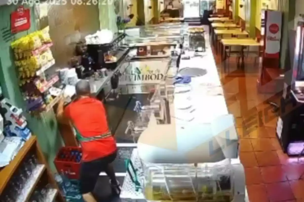 Un home amb samarreta vermella s’inclina sobre el taulell d’una cafeteria mentre una persona darrere del taulell l’observa, en un local amb taules alineades al fons. Un home amb samarreta vermella s’inclina sobre el taulell d’una cafeteria mentre una persona darrere del taulell l’observa, en un local amb taules alineades al fons.