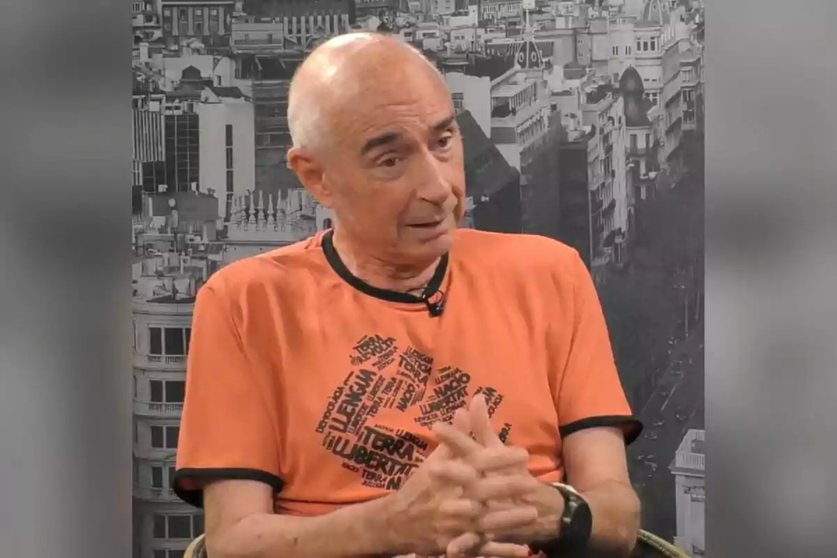 Home calb amb samarreta taronja amb paraules impreses en negre assegut amb les mans entrellaçades davant d’un fons urbà en blanc i negre