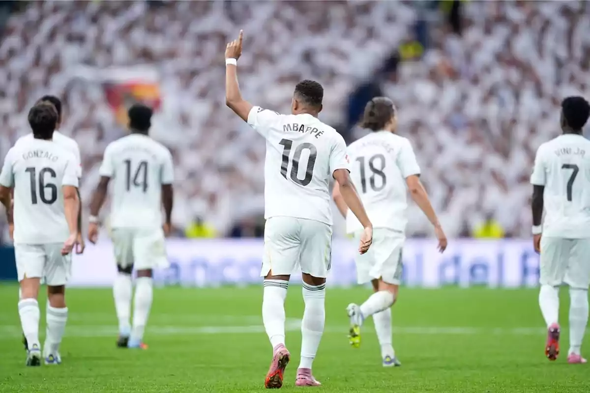 Jugadors de futbol amb uniforme blanc celebren al camp mentre un d’ells aixeca el braç assenyalant cap amunt.