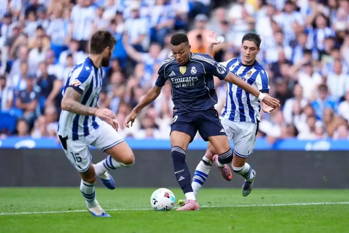 Un jugador del Reial Madrid disputa la pilota amb dos jugadors de la Reial Societat durant un partit de futbol en un estadi ple d’aficionats.