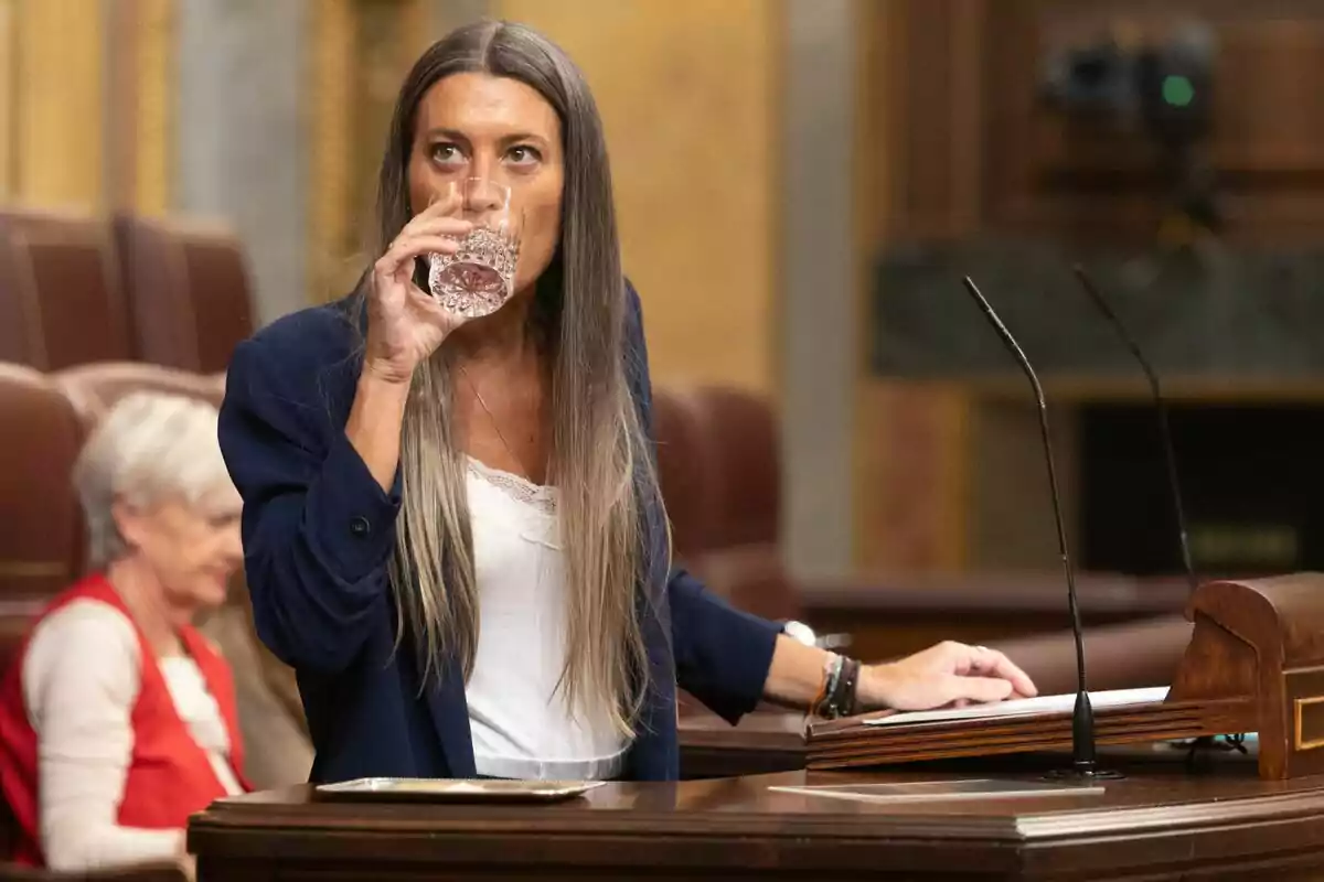 Mujer de cabello largo y lacio bebiendo agua en un vaso mientras está de pie junto a un atril en un recinto parlamentario