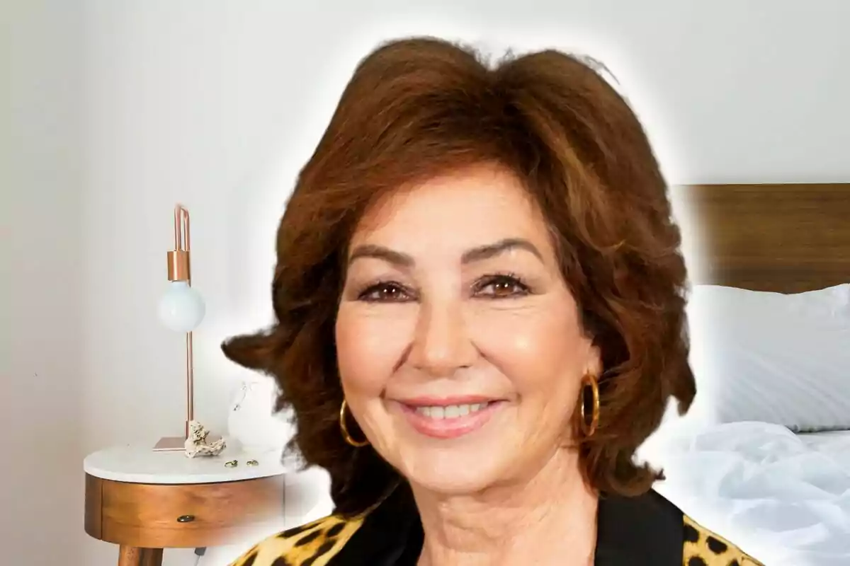 Primer pla d’Ana Rosa Quintana somrient amb arracades daurades en una habitació amb llit i tauleta de nit de fusta.