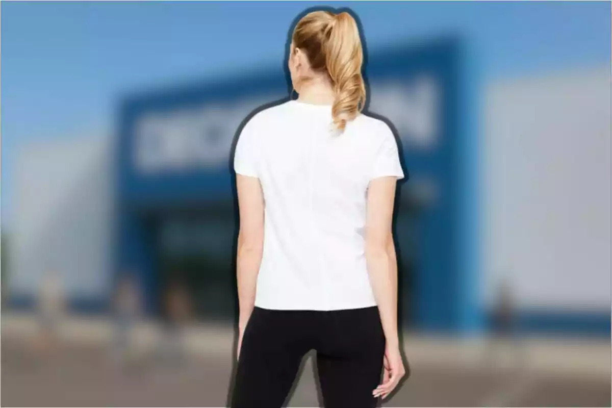 Mujer de espaldas con camiseta blanca y pantalón negro frente a un edificio azul desenfocado