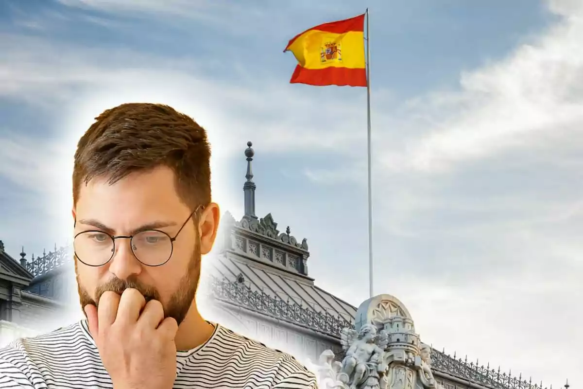 Home pensatiu amb ulleres davant d’un edifici històric i una bandera d’Espanya onejant al cel.