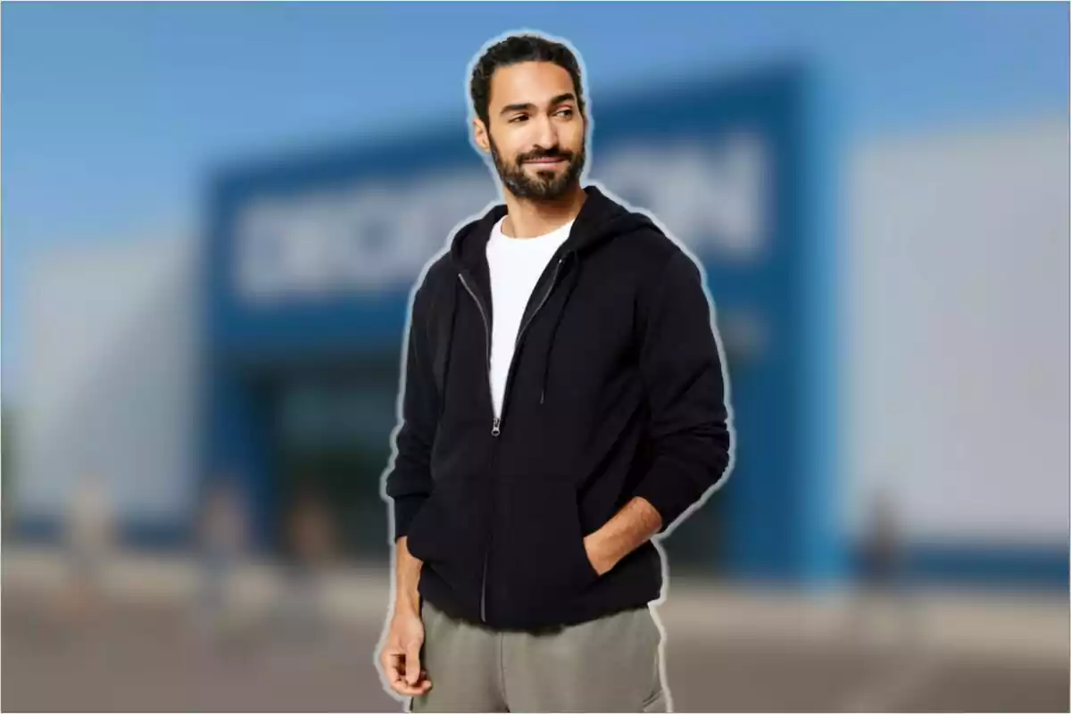Hombre joven con barba y cabello recogido viste sudadera negra y camiseta blanca de pie frente a una tienda desenfocada