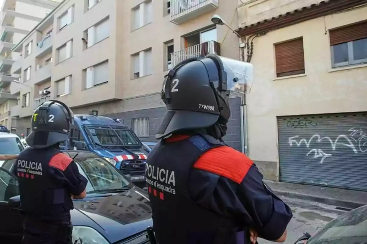 Dos agentes de policía con casco y chaleco observan una calle urbana con edificios de apartamentos y una furgoneta policial estacionada. Dos agentes de policía con casco y chaleco observan una calle urbana con edificios de apartamentos y una furgoneta policial estacionada.