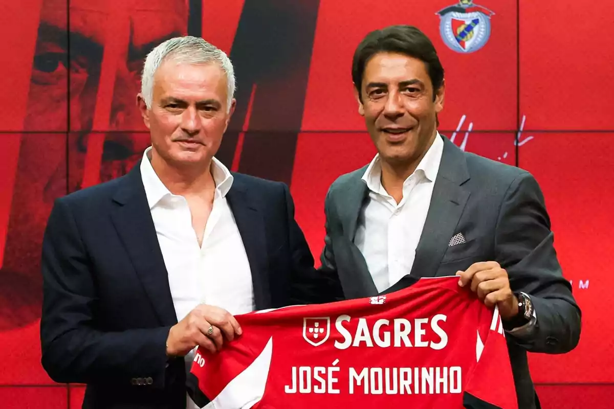 Dos homes amb vestit posen junts sostenint una samarreta de futbol vermella amb el nom de José Mourinho sobre un fons vermell amb l’escut d’un club esportiu.